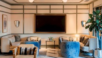 A modern, elegant hotel lounge with beige tone walls, tufted sofas, blue chairs, a large wall TV, and a potted plant by the window, cozy lighting.