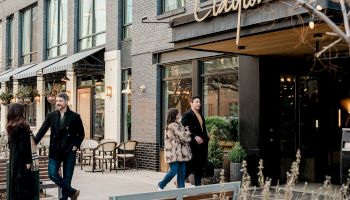 People stroll outside a chic storefront, chatting near a cafe seating area by a modern brick building with glass windows, stylish signage, and plants.