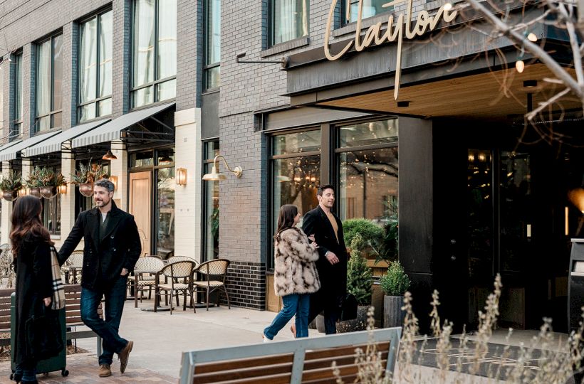 People stroll outside a chic storefront, chatting near a cafe seating area by a modern brick building with glass windows, stylish signage, and plants.