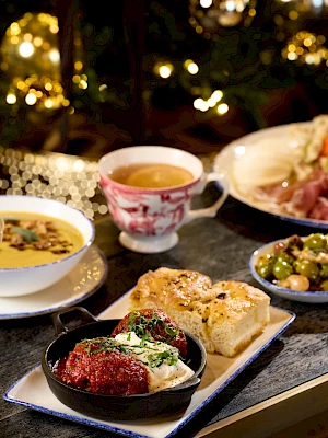A cozy, festive table with a spread of appetizers and drinks including soup, bread, meats, dips, a pink cocktail, and ambient fairy lights in the background.
