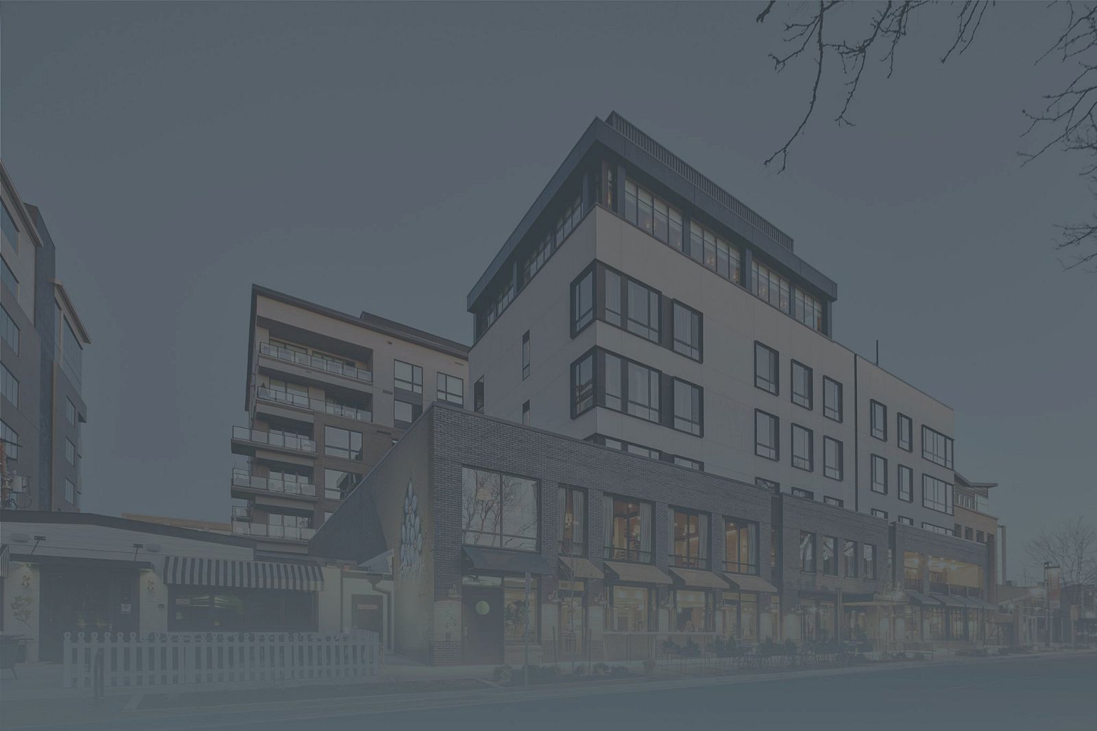 A modern multi-story building complex with large windows and a mixed-use design, set along a street with some trees.