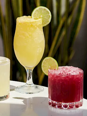 A tray of colorful cocktails: a tall yellow drink garnished with a lime wheel, a bright red/berry cocktail, and a pale yellow one in the background.