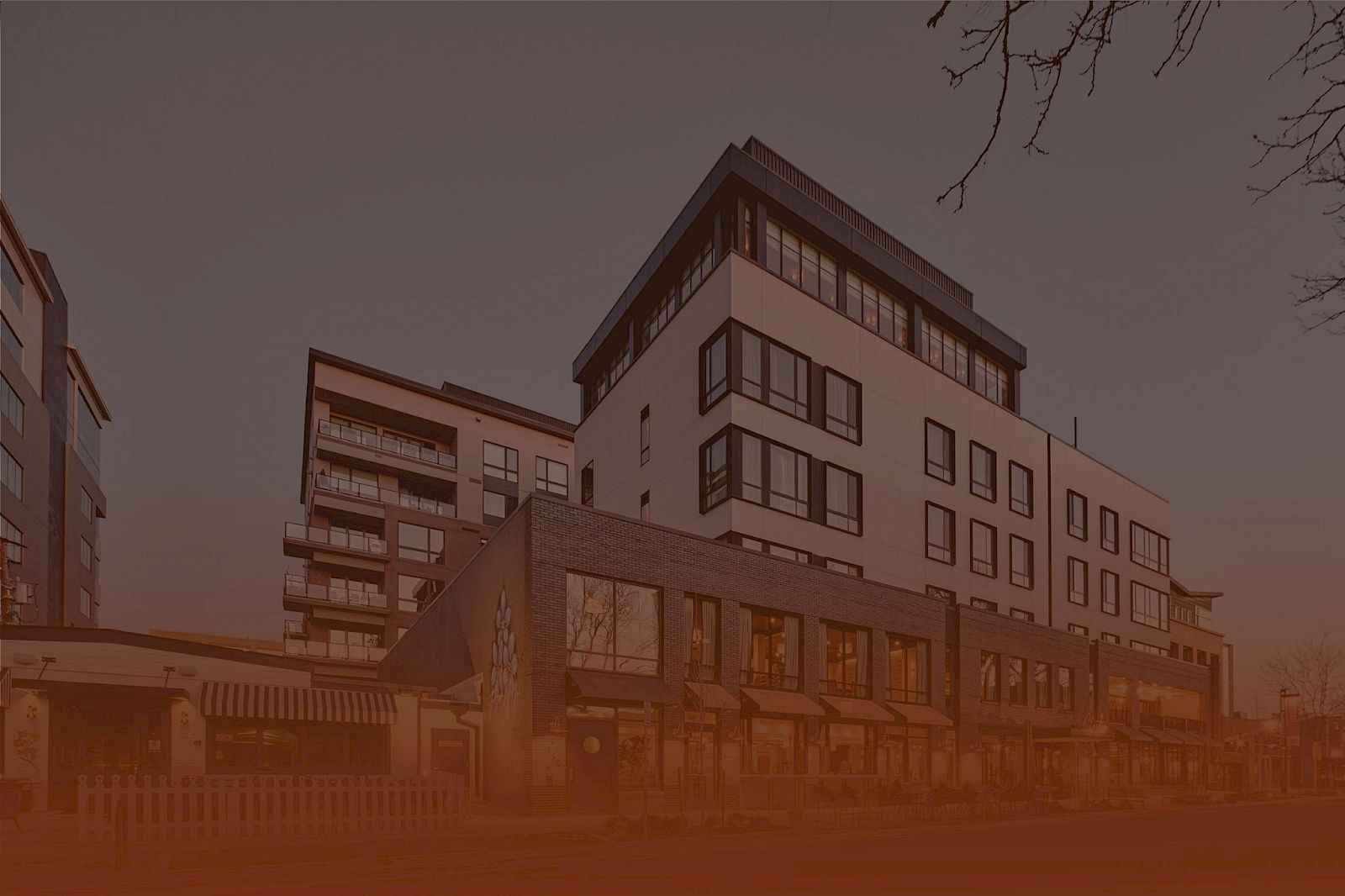 A modern multi-story building complex with large windows, brick and white fa&ccedil;ade, set against a dusky sky.