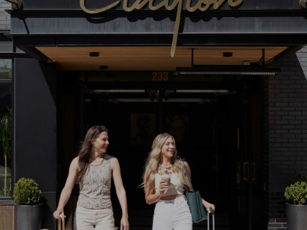 Two stylish women exit a city hotel with rolling luggage and a coffee, heading into a doorway under a sign that reads &ldquo;Clayton.&rdquo;
