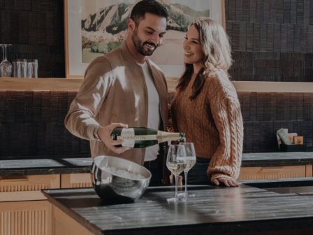 A couple is in a kitchen, smiling as a man pours wine into glasses on a counter.