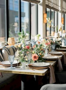 A stylish restaurant interior with long tables set for dining, chic chairs, warm lighting, plants, and a bright, inviting atmosphere.