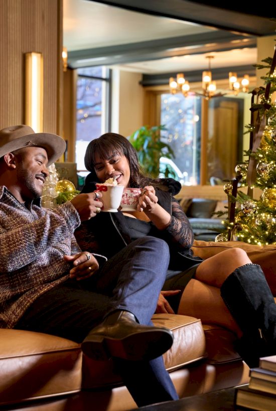 Two friends sit on a cozy sofa by a festive Christmas tree, sharing drinks and laughter in a warmly lit, stylish lounge.