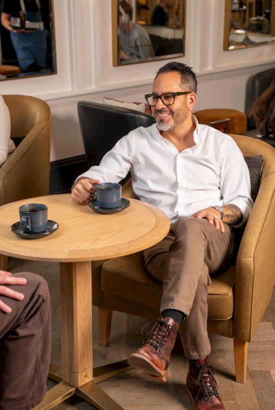 A man in a white shirt sits on a tan chair, smiling, with a mug on a wooden table in a cozy cafe. End with a period.
