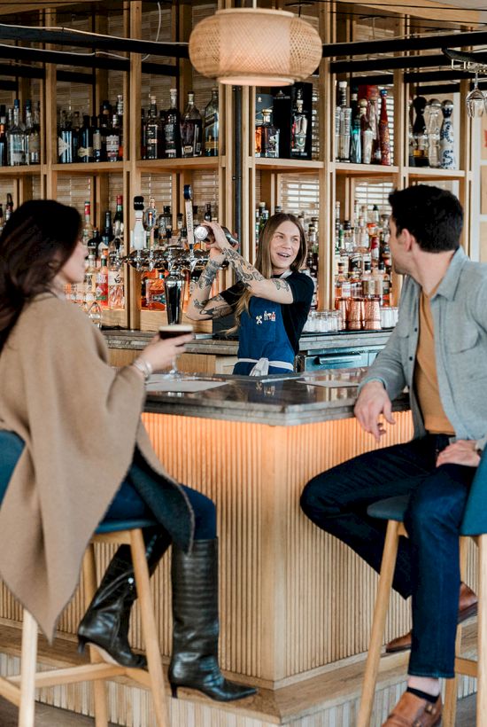 A stylish bar scene with five people chatting and sipping drinks around a curved, well-lit wooden bar with blue stools.