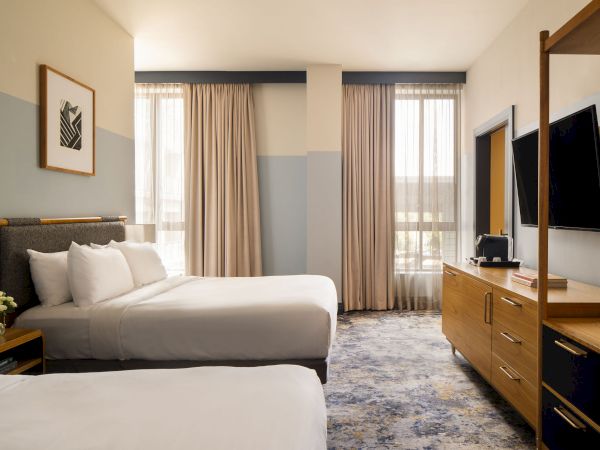 A modern hotel room with two queen beds, soft lighting, a dresser with a TV, floor-to-ceiling windows, and neutral decor with blue-gray walls.