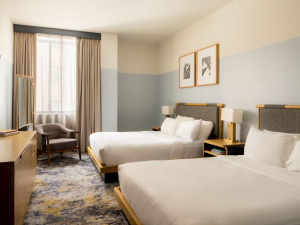 Two queen beds in a modern hotel room, with a seating area by a window, soft blue walls, and framed art above the beds.