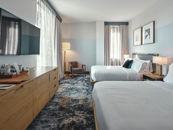 Two hotel beds with white linens, a dresser and TV on the left, a seating chair by a lamp, soft blue wall accents, and framed art above the beds.