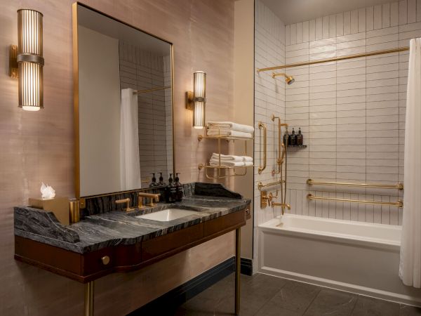 A luxurious bathroom with a double vanity, dark counter, wall mirror, warm lighting, and a bathtub/shower combo with white subway tiles and gold accents.