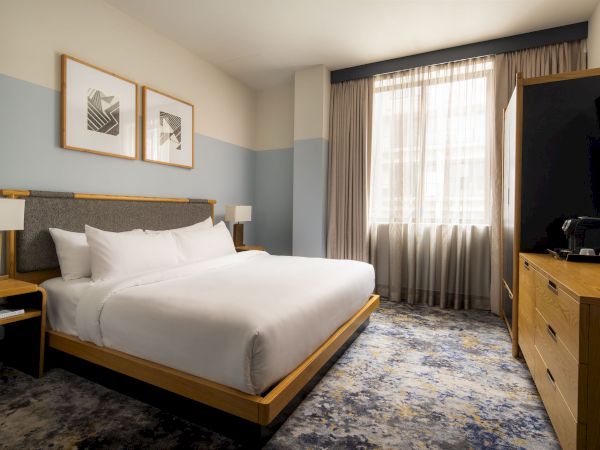 A modern hotel room with a king bed, light wood furniture, blue-gray walls, framed art, a large window with sheer curtains, and a flat-screen TV.