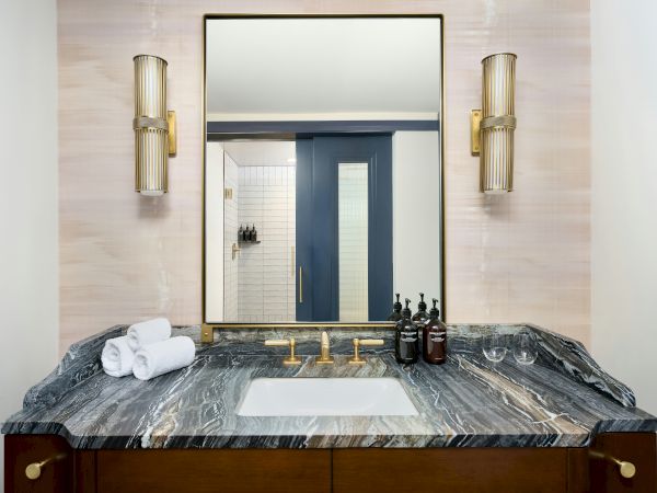A bathroom vanity with a dark marble counter, a white sink, a mirror, two gold sconces, rolled towels, and dark wood cabinets.