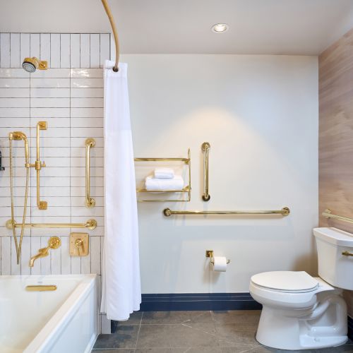 The image shows a bathroom with a bathtub, shower, toilet, and gold-colored fixtures and grab bars for accessibility and safety.