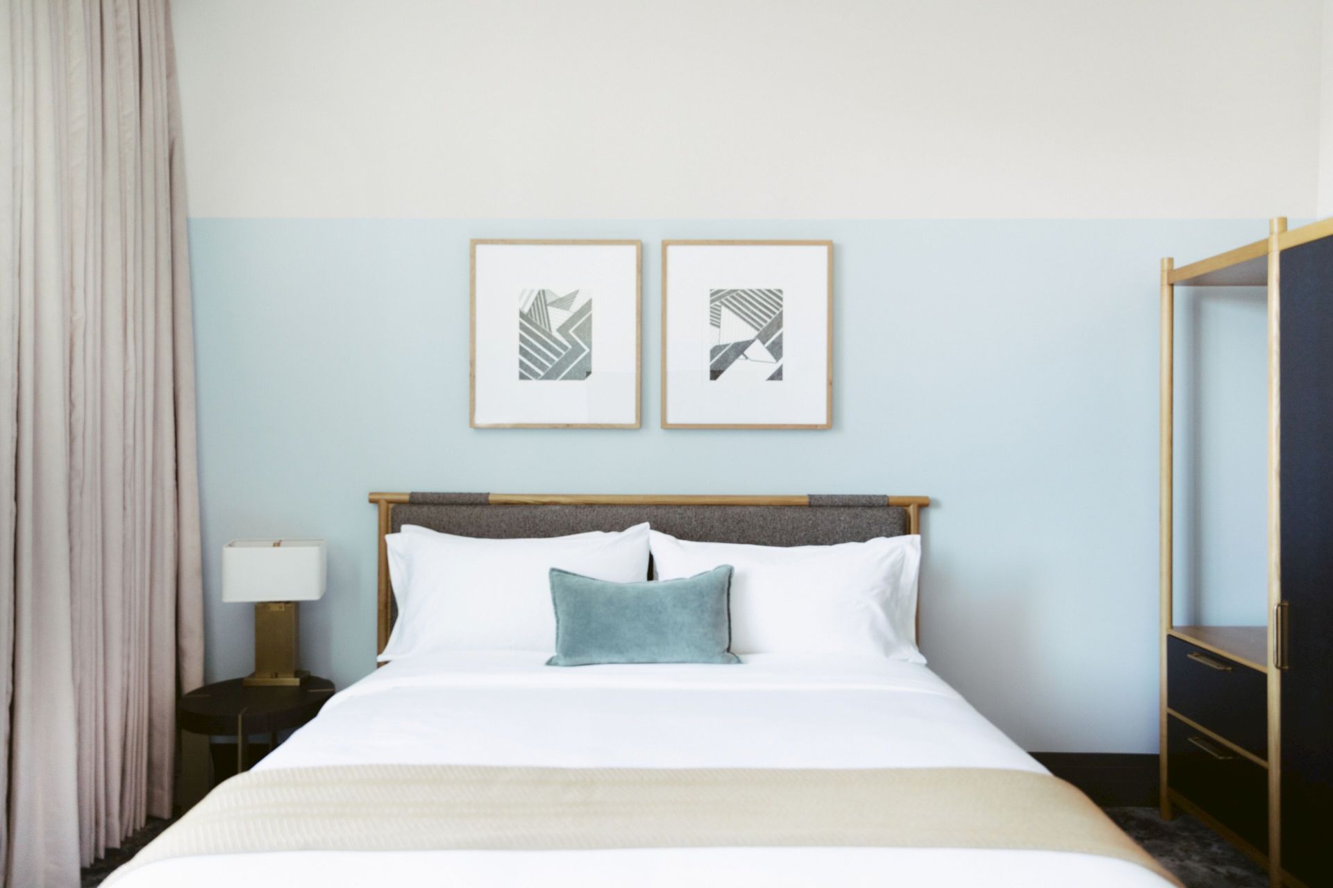 A neatly made bed with white linens, a light blue pillow, and a headboard, flanked by a lamp, artwork, and an open wardrobe.