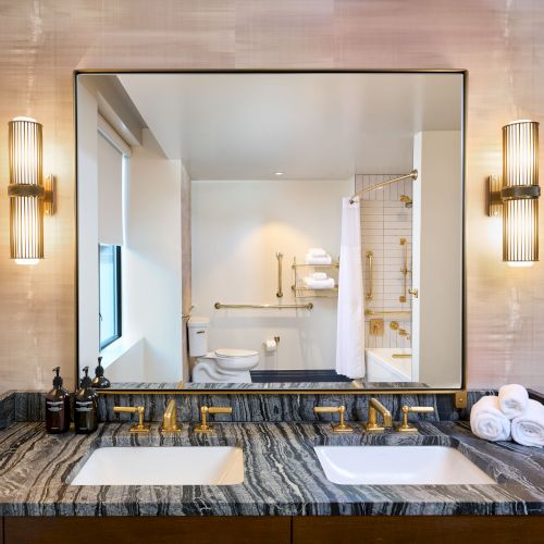 The image shows a bathroom with a large mirror, twin sinks on a black marble counter, elegant lighting, and neatly arranged toiletries.