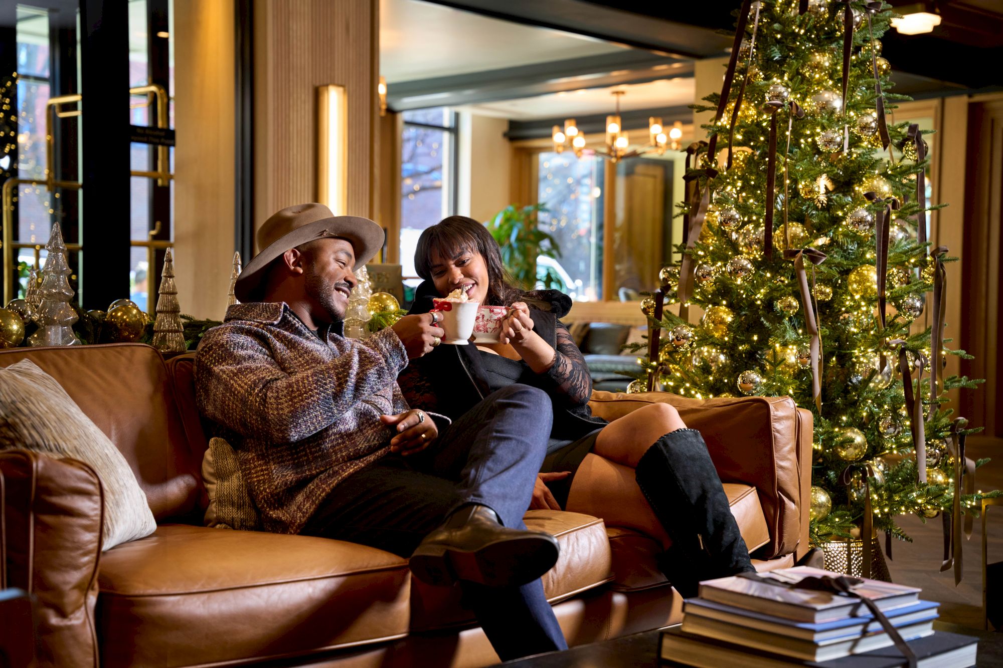 A couple relaxes on a couch, sipping cocoa beside a glowing Christmas tree.