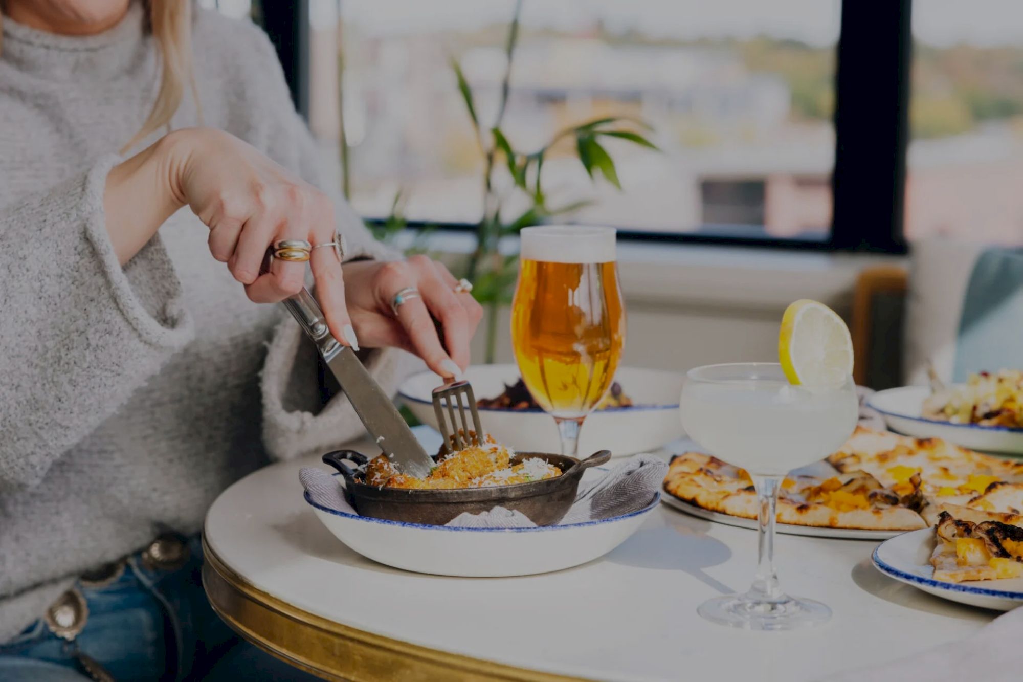 A person is dining, with a fork and knife, enjoying a meal. Drinks, including a lemon-garnished cocktail, are on the table.