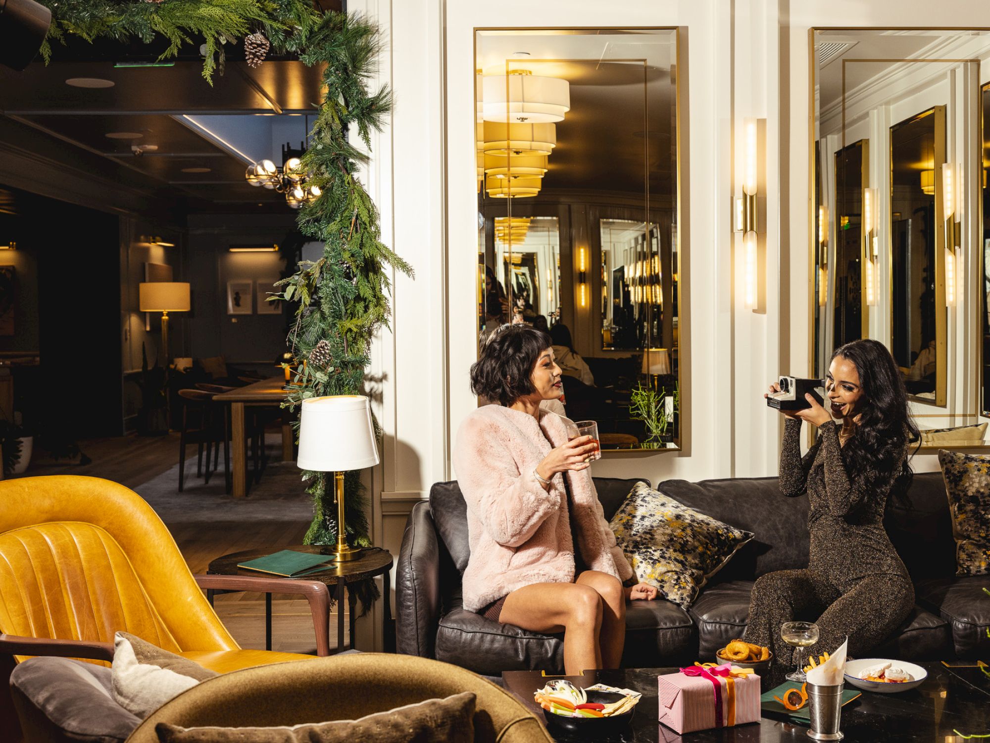 Two people are sitting on a couch, enjoying drinks in a cozy, decorated room with warm lighting and festive greenery.