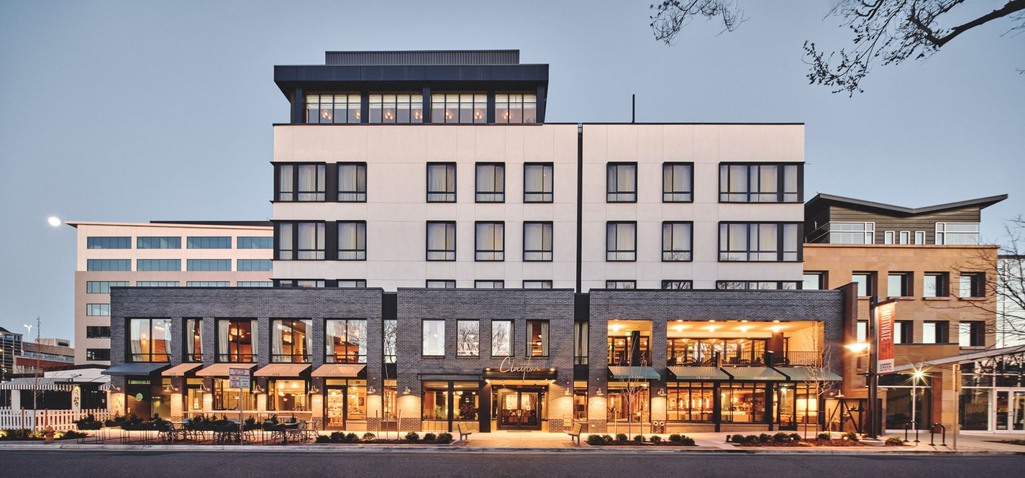 The image shows a modern multi-story building with various shops and businesses on the ground level, featuring large windows and a sleek design.