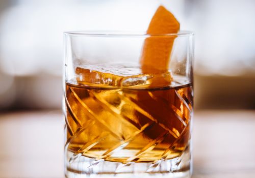 A glass of amber-colored liquid with ice and an orange peel garnish rests on a polished surface.