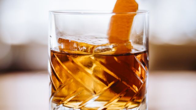 A glass of amber-colored liquid with ice and an orange peel garnish rests on a polished surface.