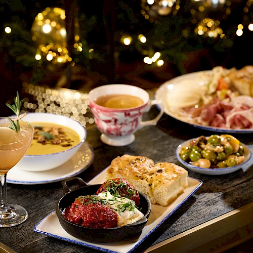 A festive table of assorted dishes: cocktails, soups, bread, roasted meats, veggie sides, and pastries, all under warm holiday lights.