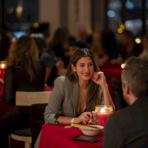 Two people dining in a cozy, candlelit restaurant with red tablecloths; a woman smiles at a man across the table.