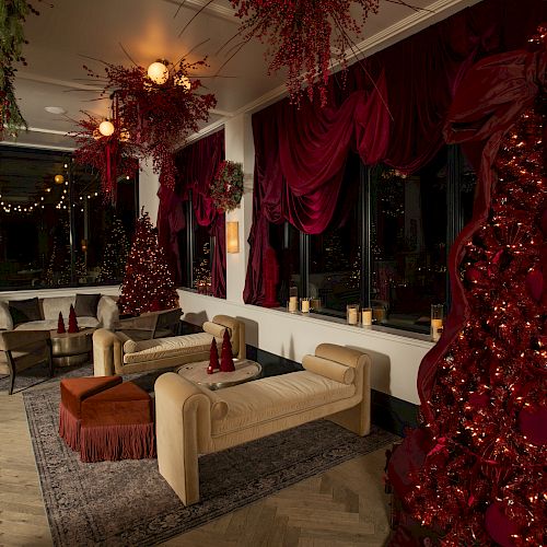 A cozy lounge with cream sofas, a red Christmas tree, hanging greenery, red drapes, and warm lighting in a festive, elegant setting.