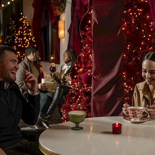 A man and a woman laugh and chat at a festive outdoor table with drinks; twinkling lights and red decorations create a warm, romantic setting.