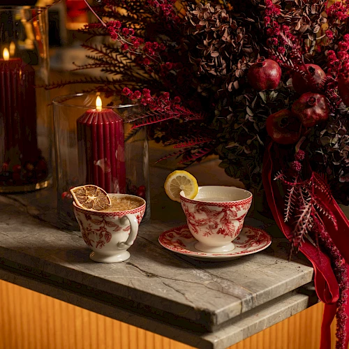 A cozy bar or dining scene with two delicate teacups on a marble counter, a lit red candle, and a lush winter floral arrangement in deep reds and purples.