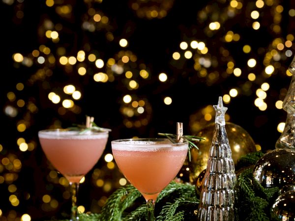 Two pink cocktails with little umbrellas on a festive, sparkly backdrop; evergreen branches and a decorative bottle add a cozy holiday vibe.