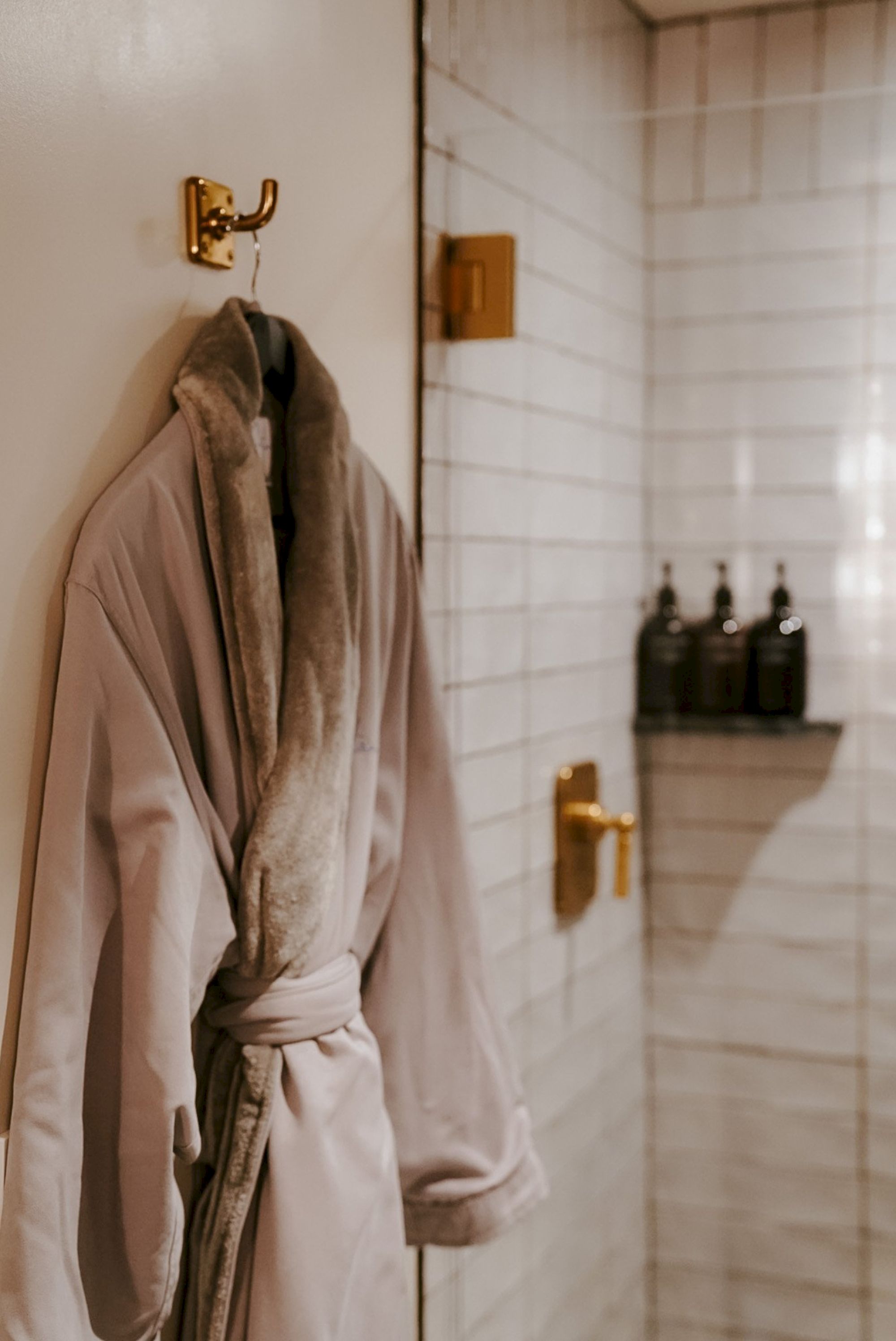 A plush beige bathrobe hangs on a hook by a door in a bright tiled bathroom, with a glass shower area and dark bottles on a shelf in the background.