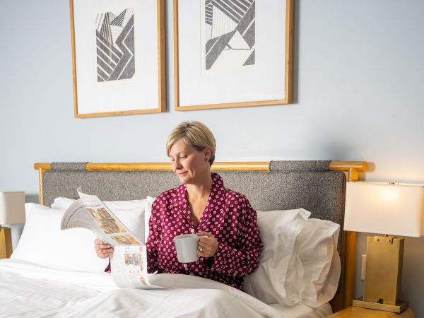 A woman in a red polka-dot shirt sits up in bed, reading a newspaper and holding a mug, with framed art above and nightstands on either side.