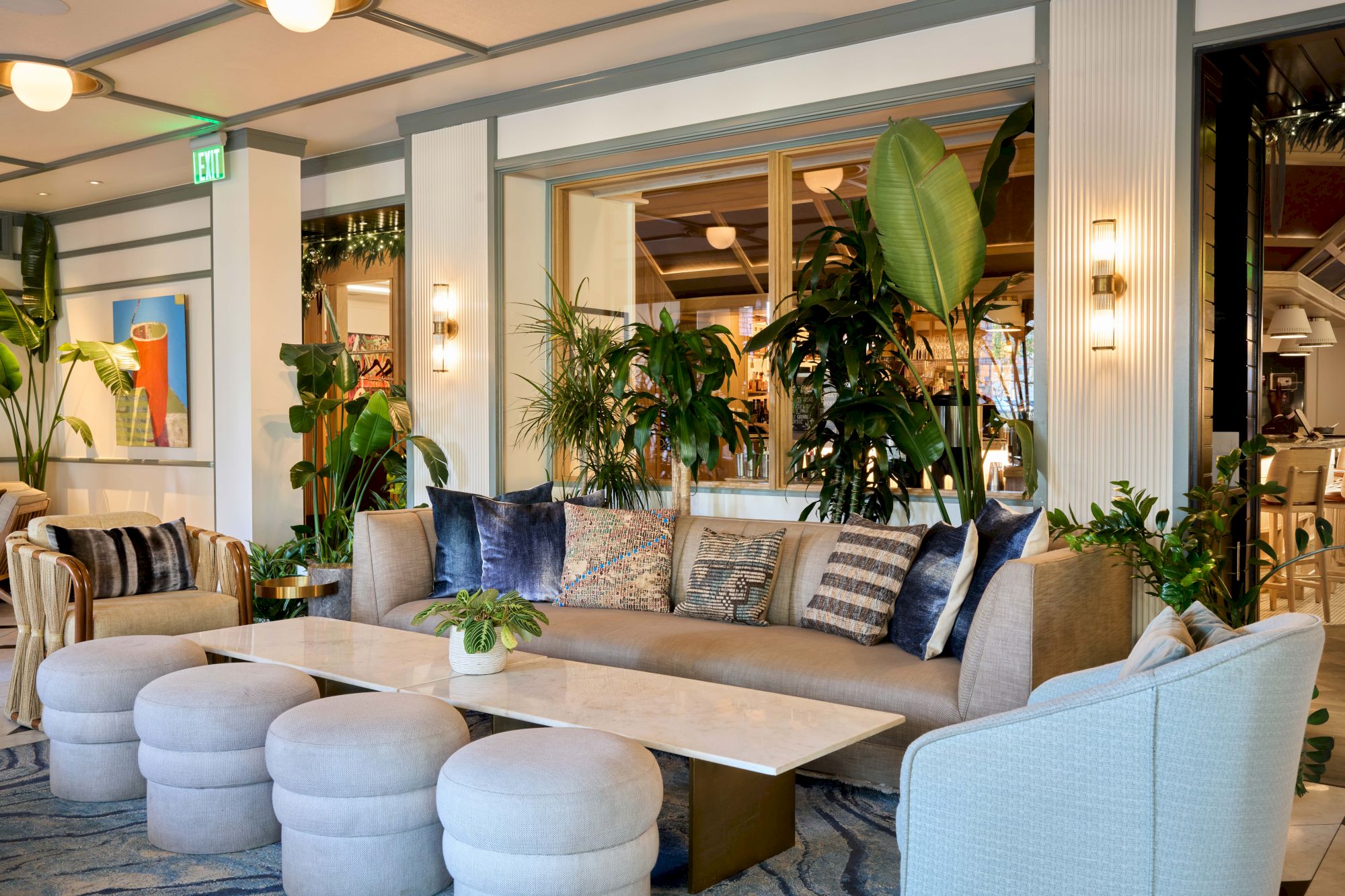 A bright, modern lounge with a curved beige sofa, multiple blue and gray cushions, white cylindrical ottomans, green plants, and warm wall lights.