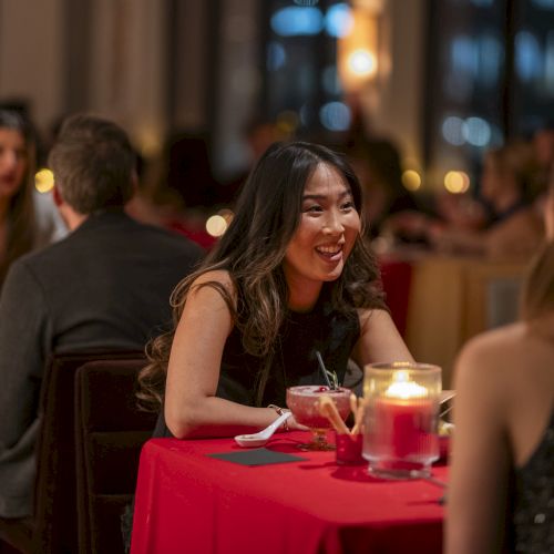 A smiling woman in a black dress chats with a friend at a candlelit dinner date; warm restaurant ambience with red tablecloths.