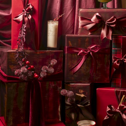 A cozy pile of red gift boxes wrapped with ribbons, topped with a few candles and cups, against a rich burgundy backdrop.