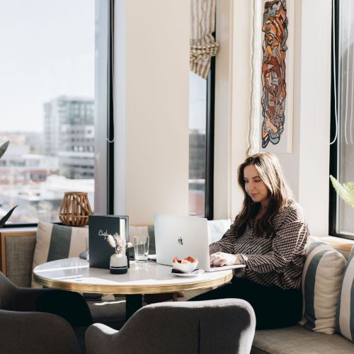 A woman sits in a bright cafe corner, laptop open, sipping coffee near a window with plants and modern decor, creating a cozy workspace.