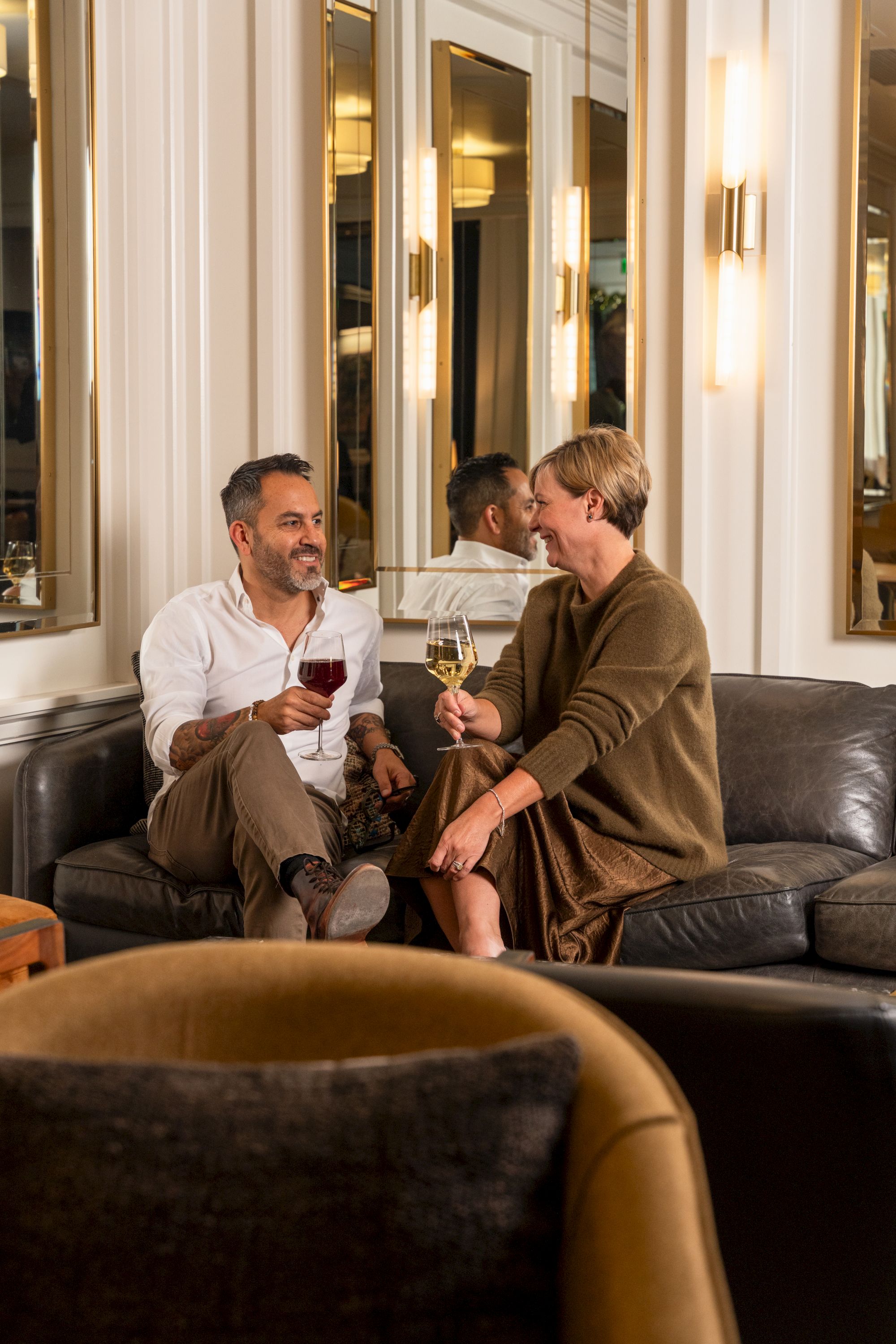 Two people sit on a plush sofa, smiling and clinking wine glasses in a stylish, mirrored lounge. A cozy, upscale vibe.