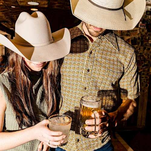Two people wearing cowboy hats are clinking beer glasses in a rustic setting, beer in hand and smiles ready to toast.