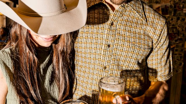 Two people wearing cowboy hats are clinking beer glasses in a rustic setting, beer in hand and smiles ready to toast.