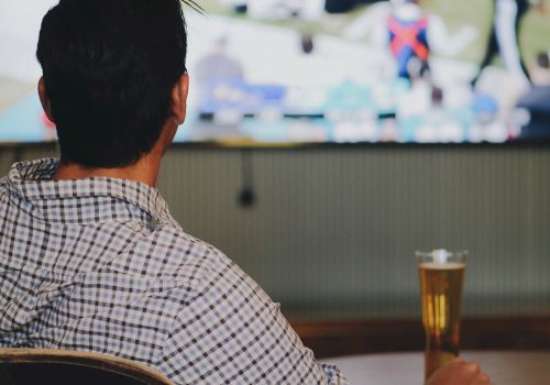A man watches a sports game on TV, seated at a bar with a beer in front, catching action on a large screen.
