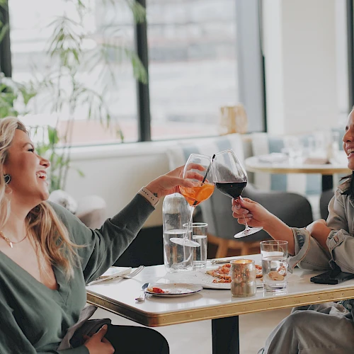 Two women share a joyful toast in a bright cafe, clinking glasses and smiling as they enjoy drinks and conversation.