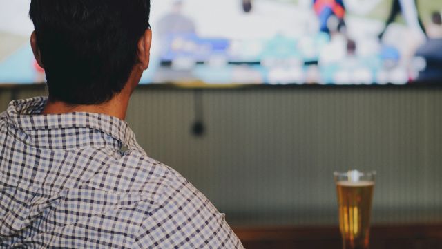 A man watches a sports game on TV at a bar, beer on the table, blurred players on screen, cozy corner vibe, focused on the action.