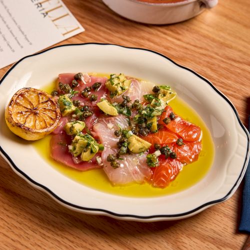 Sliced cured meat and vegetables on a yellow oil drizzle, adorned with herbs, citrus wheel, and a pasta-like topping on a white plate.