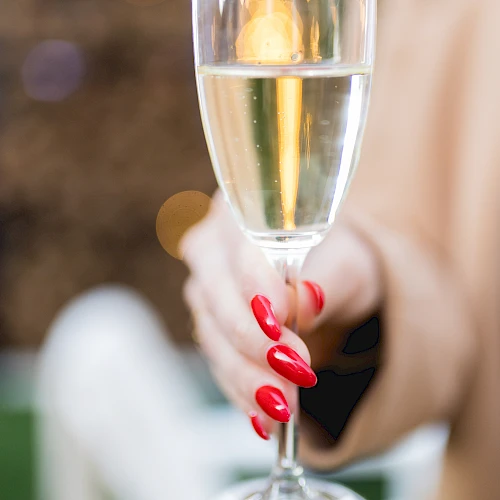 A close-up of a champagne flute held by a person with red nail polish, blurred background, sparkling wine inside the glass, and a soft bokeh.