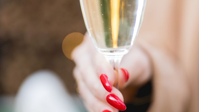 A close-up of a champagne flute held by a person with red nail polish, blurred background, sparkling wine inside the glass, and a soft bokeh.