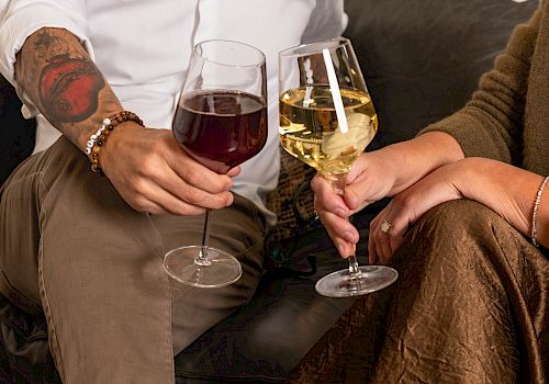 Two people clink wine glasses on a cozy couch, one wearing brown pants and boots, the other in a rust dress; relaxed, intimate cheers.